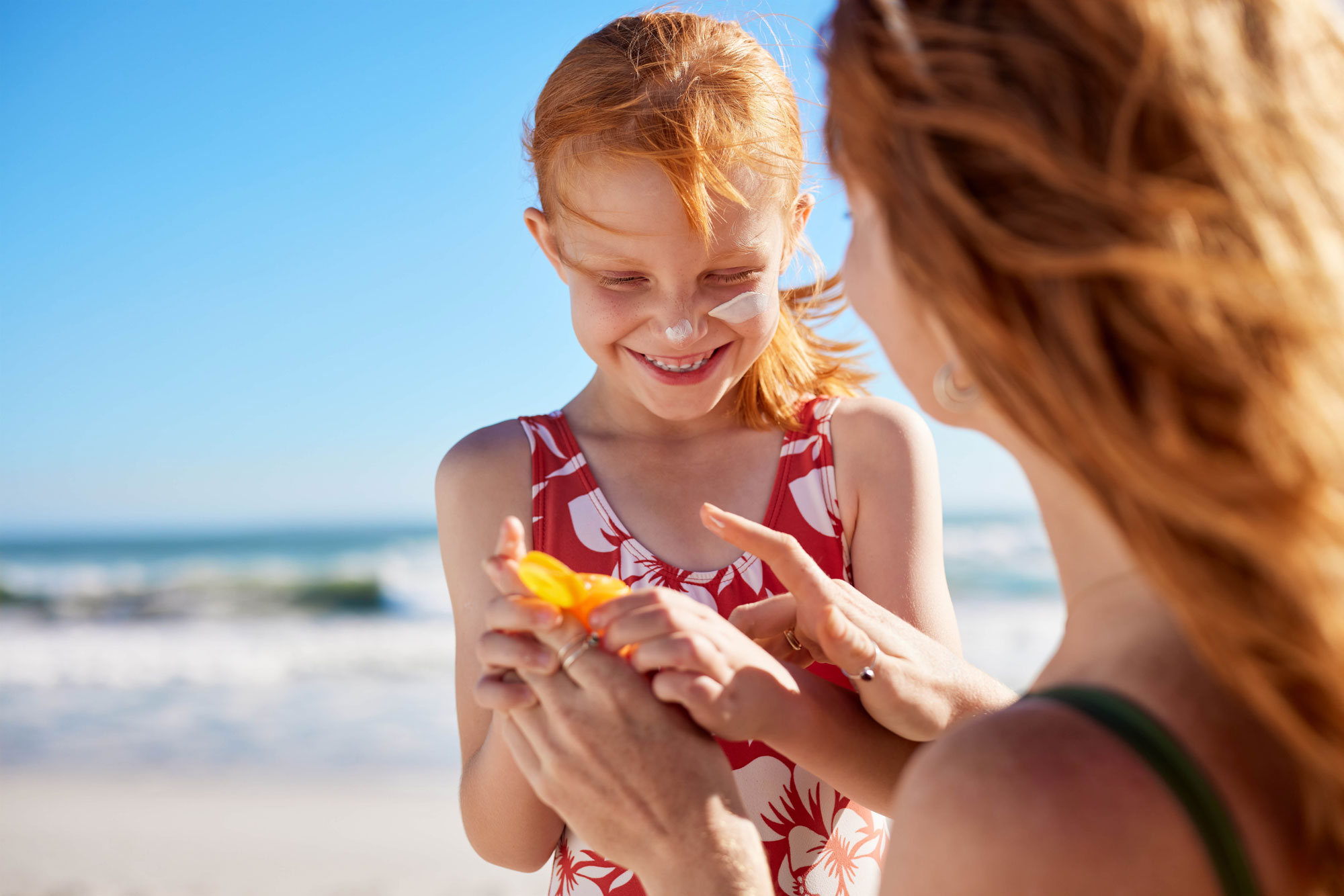 A woman with curly red hair shot from behind. she is applying sunscreen to a young white girl who also has red hair. They are at the beach. The young girl is smiling and having a fun day.