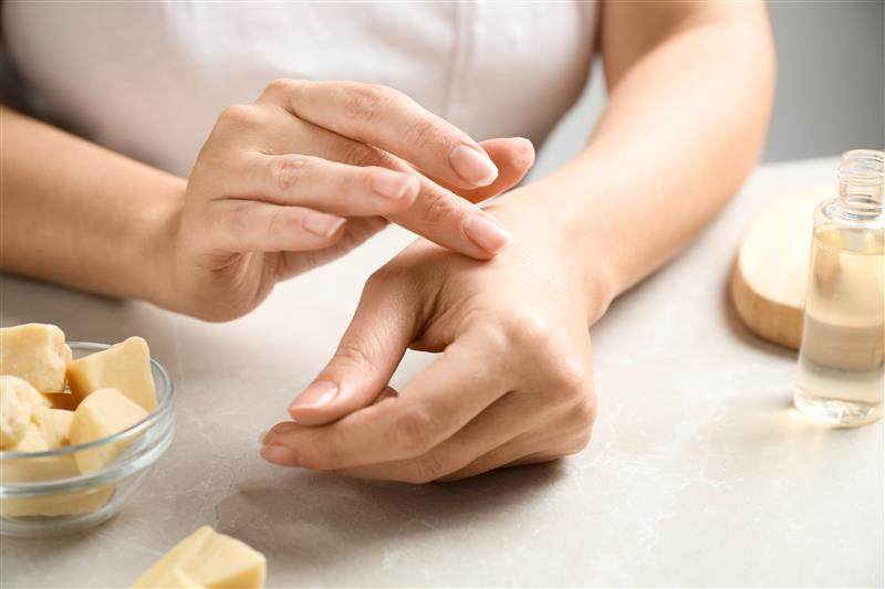 Close-up on a White woman's hands. She is sitting at a table. At the table is some cocoa butter. She is applying the cocoa butter to her hands.