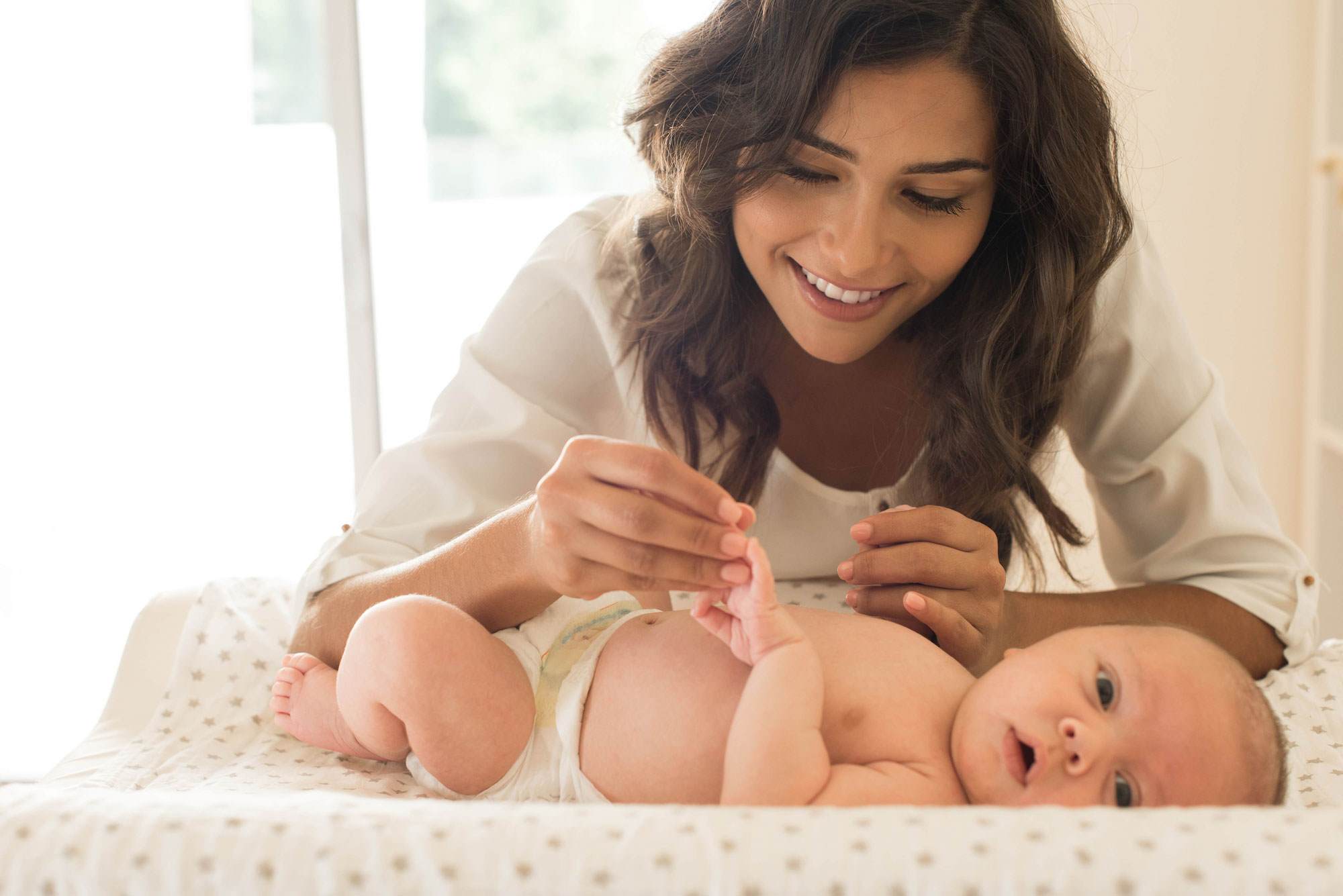 Young white woman with long brown straight hair playing with a baby who is lying on a changing mat,