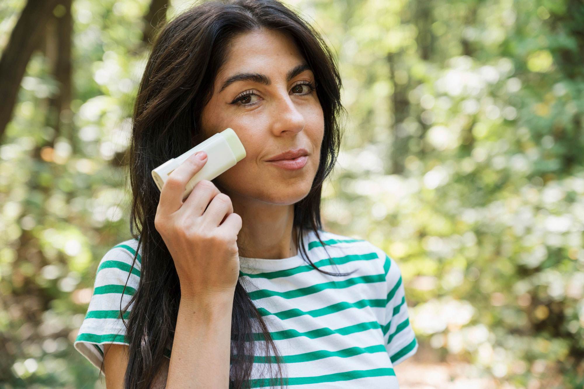 Young white woman with long black hair holding up a foundation stick and applying it to her face.