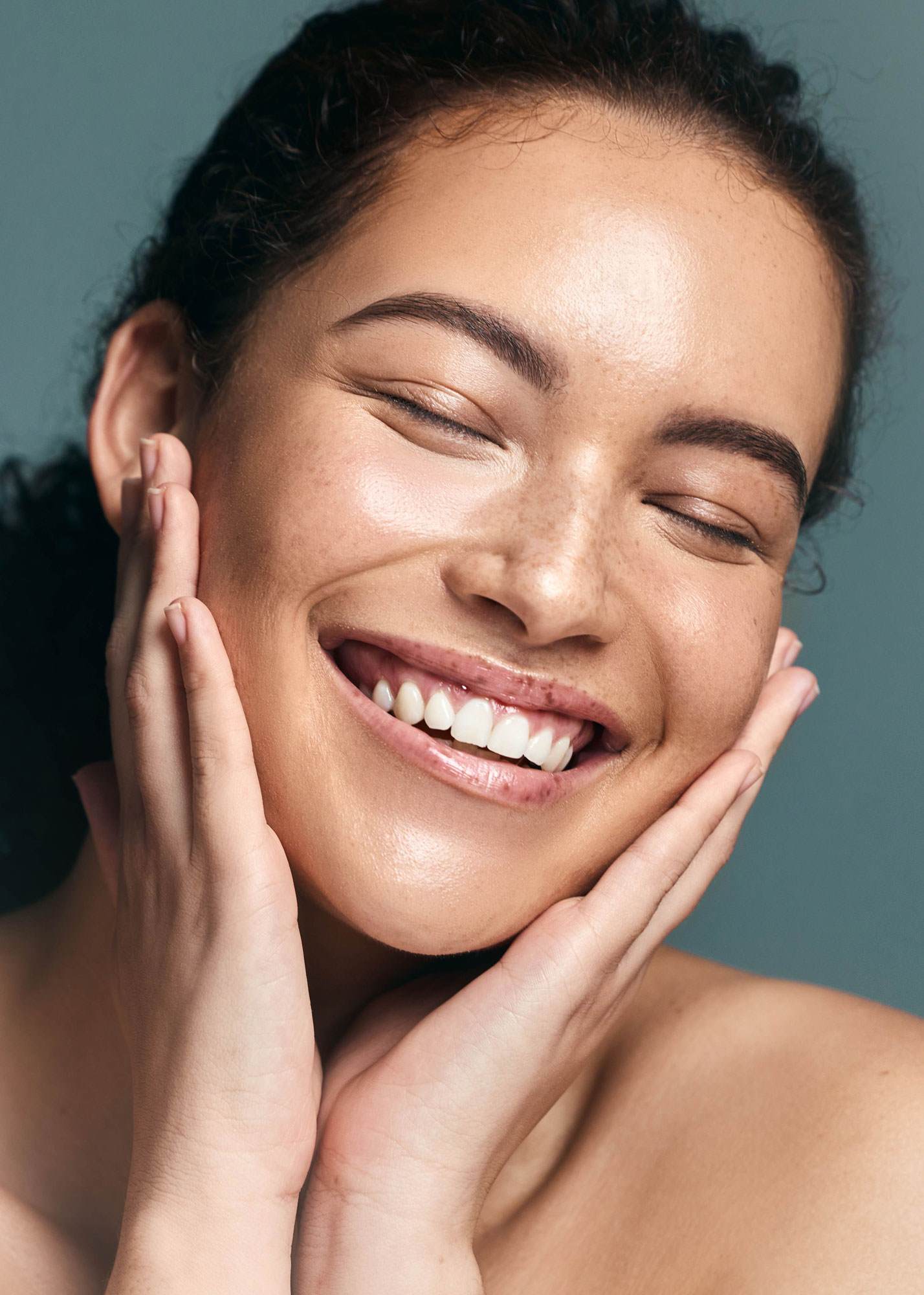 Young woman with light brown skin and brown hair smiling while holding her hands to her face.