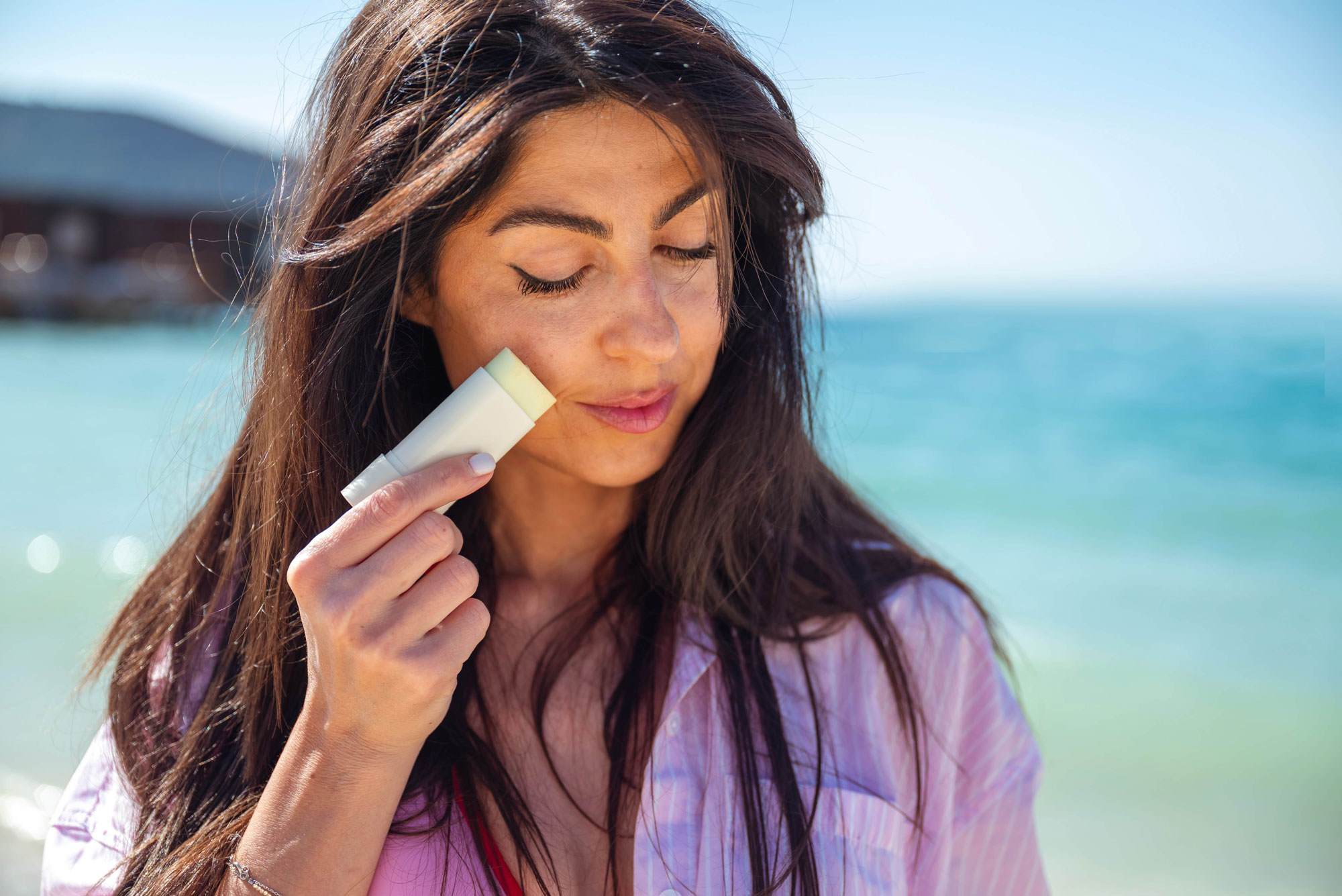 Young white woman with long straight brown hair applying a sunscreen stick to her face.