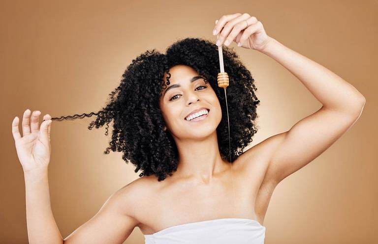 Young woman with brown skin and brown naturally curly hair smiling and looking at the camera while she teases her hair with one hand and drizzles honey with the other.