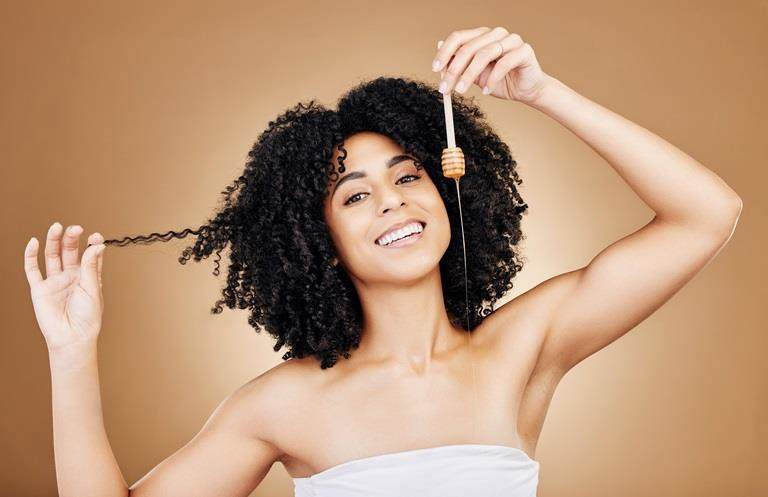 Young woman with brown skin and brown naturally curly hair smiling and looking at the camera while she teases her hair with one hand and drizzles honey with the other.