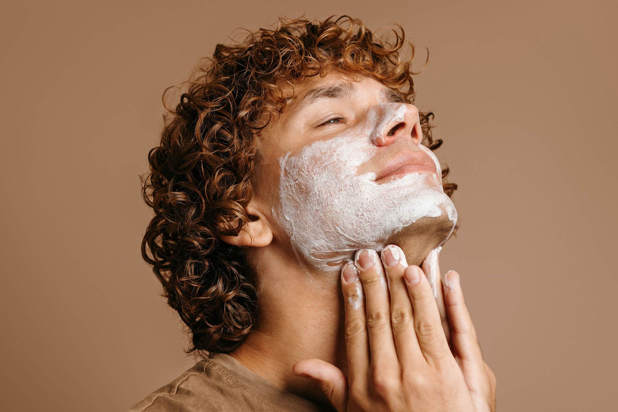 One young white man with curly red hair applying gentle facial cleansing gel to his face. The gel has lathered up into a white foam. His eyes are closed and he looks to be enjoying the sensation.