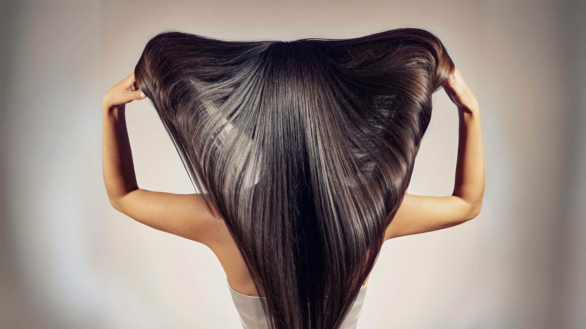 Photo of a woman from behind. She has olive skin and long brown hair. She is lifting her hair with her arms, spreading it and showing how long, healthy, and shiny her hair is.