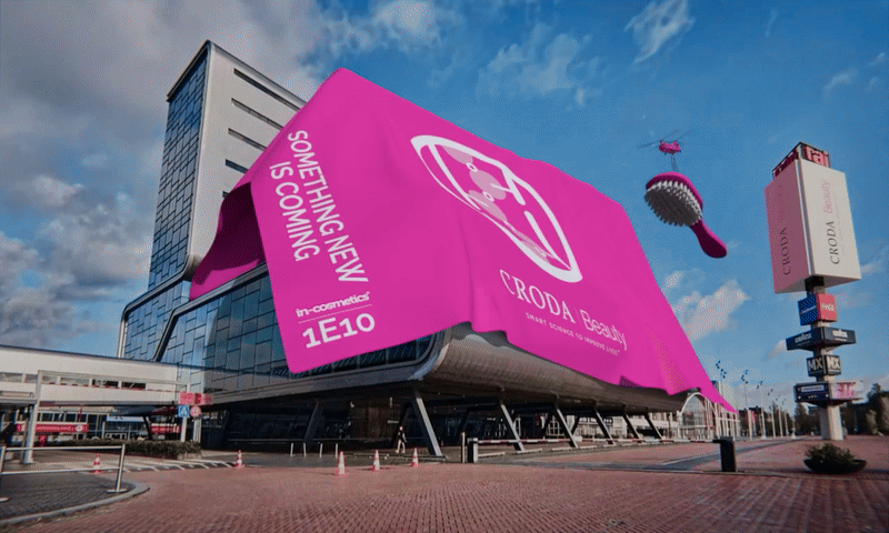 A large pink tarpaulin covering the RAI Amsterdam building. Written on one side are the words "something new is coming". A helicopter flies overhead carrying a giant hairbrush.