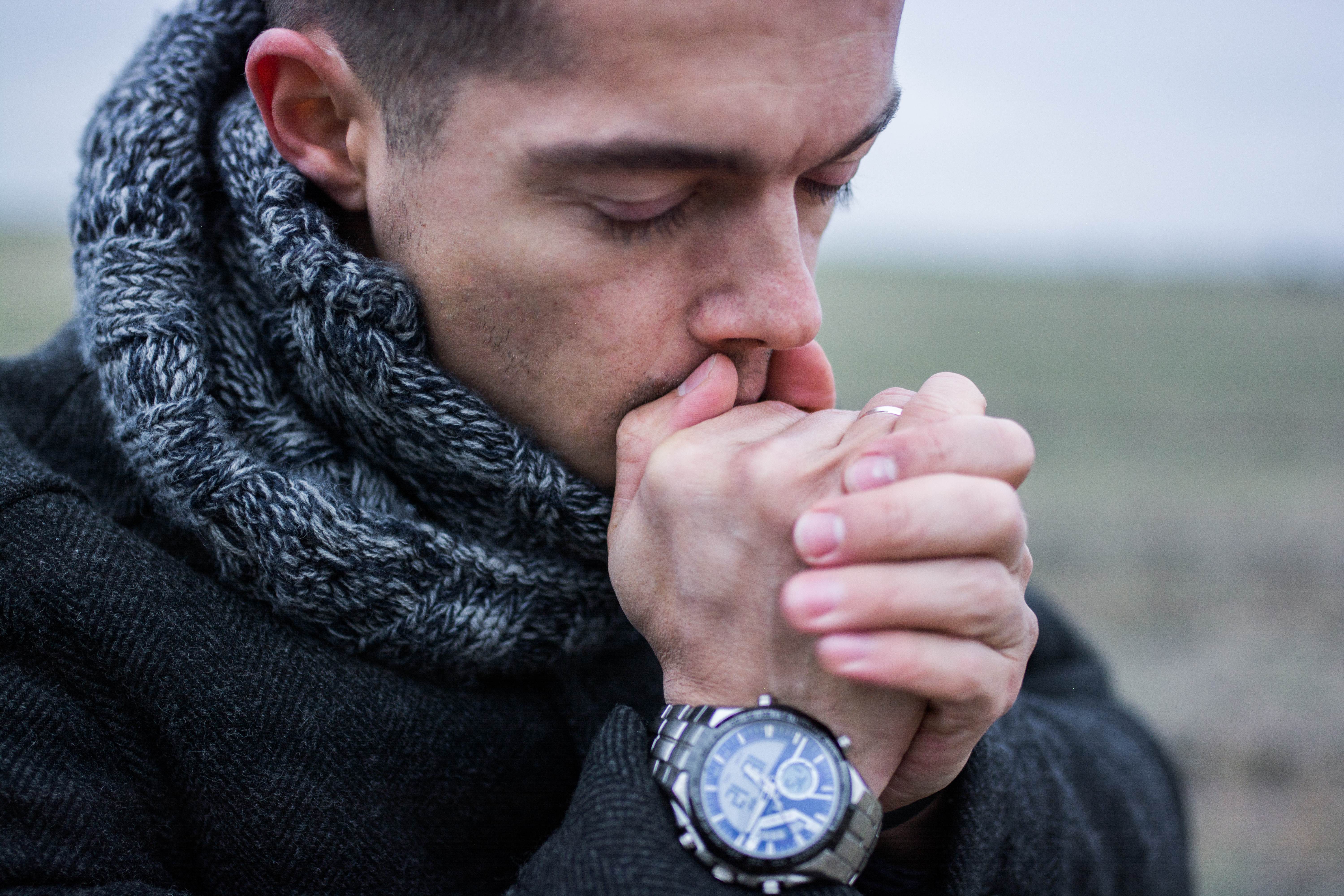 A young man with dark hair blowing on his hands to keep them warm against a cold countryside backdrop