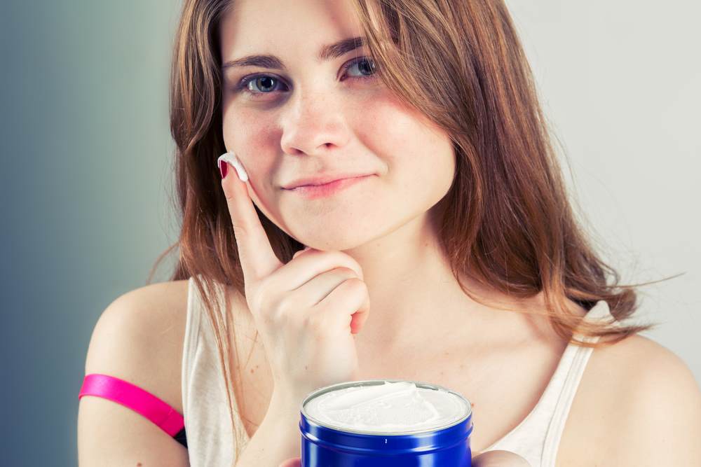 Young white woman with red hair and freckles smiling at the camera and applying  a white cream to her face