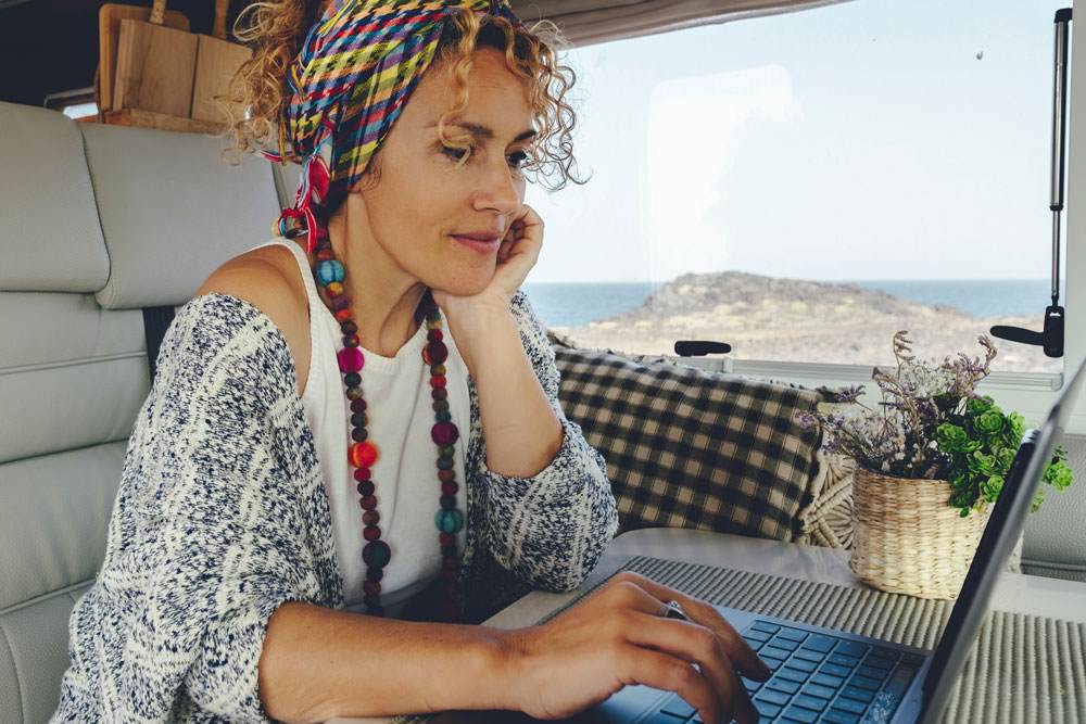 White woman in her 30s with curly light brown hair tried up in a headscarf. She is sitting in a caravan and working on a laptop.