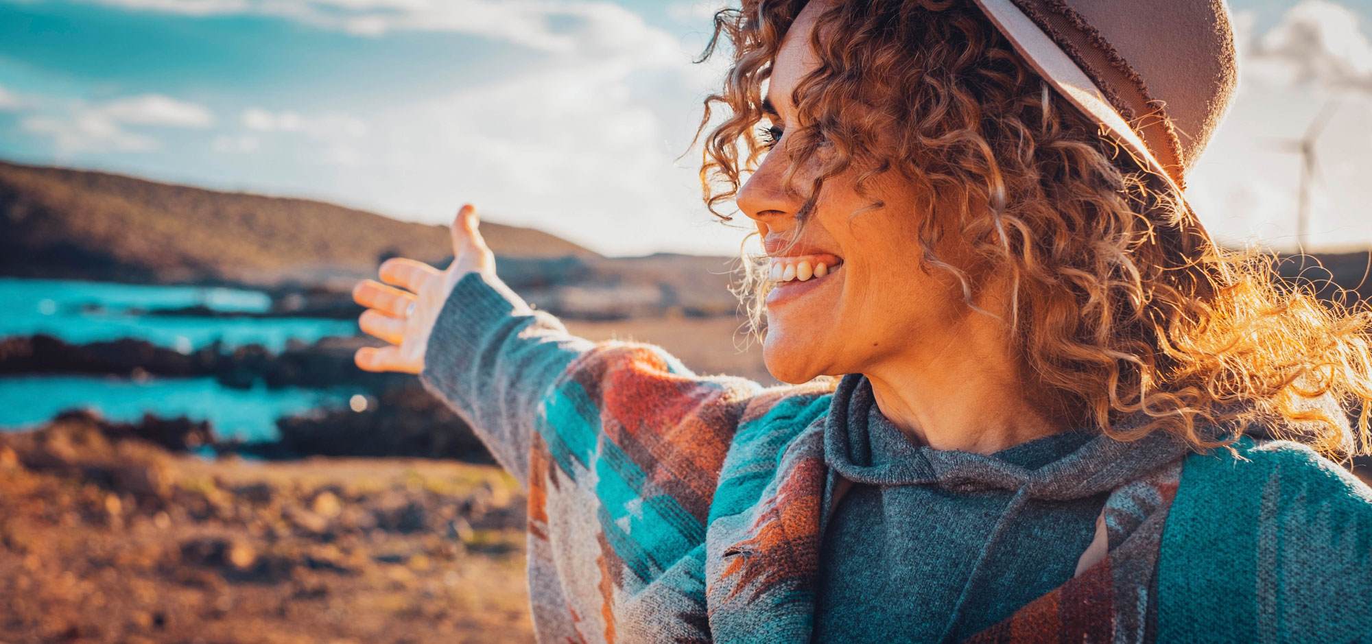Middle aged woman with brown curly hair and light brown skin smiling and looking to the left. She is in the desert with the sun shining on her. She is wearing a brown hat and blue poncho.