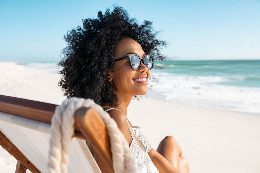 Young black woman with curly black hair in a natural style. She is wearing sunglasses, smiling and sitting on a beach chair.