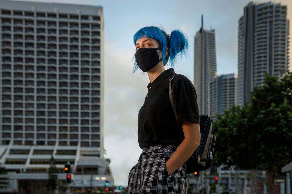 Young white woman with blue straight hair. She is wearing a black facemask and standing in a city.