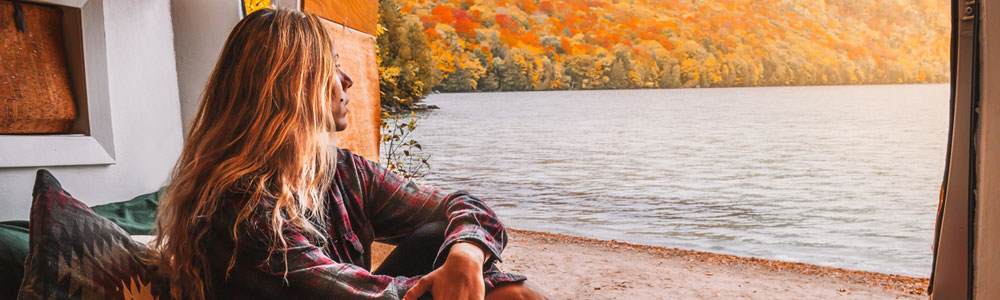 Young white woman with light brown straight hair. Her face is mostly obscured but we can see she is looking to the right out over a beautiful autumnal lake.