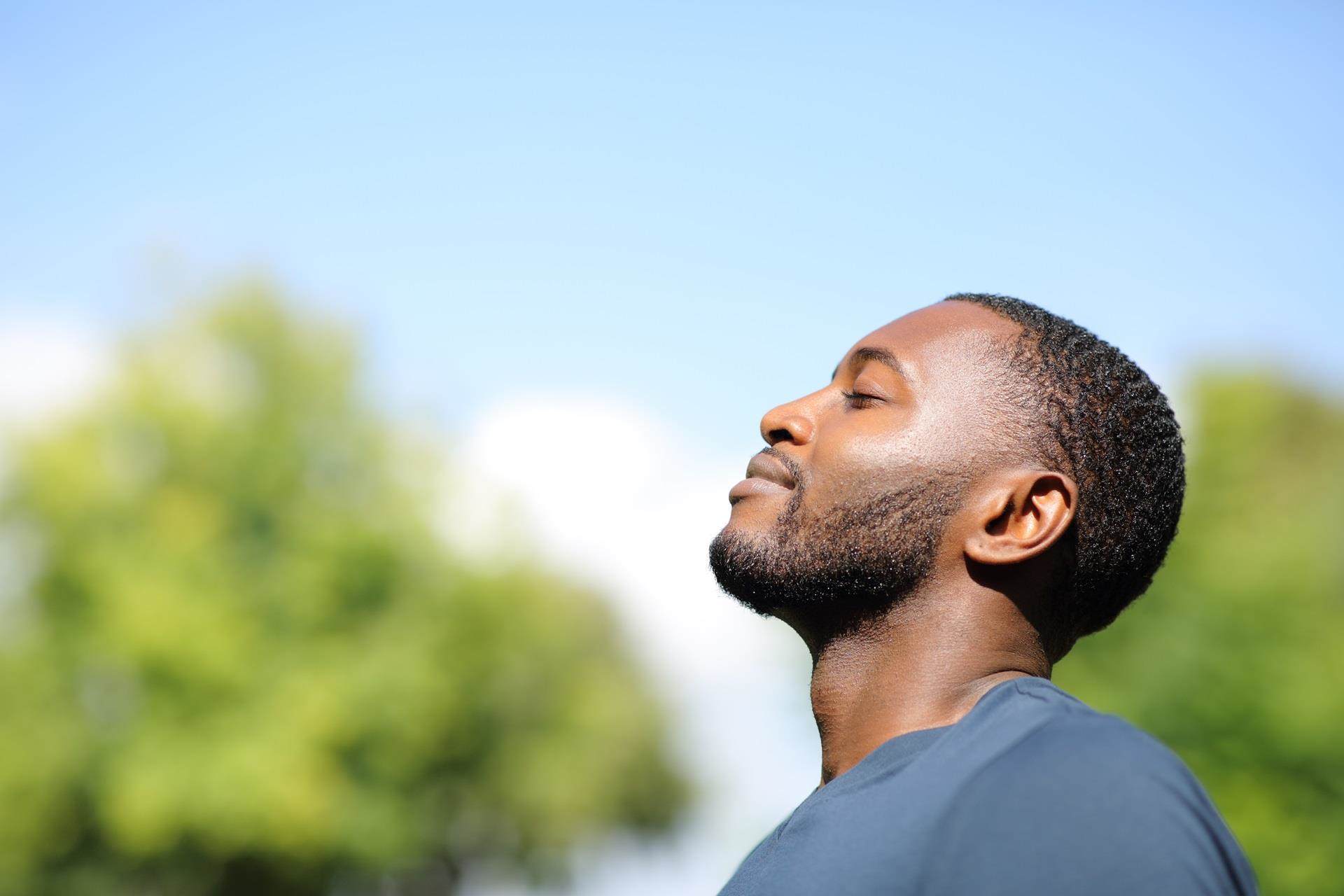 Young black man with closely shaved black hair and beard, smiling with his eyes closed in the sunshine.