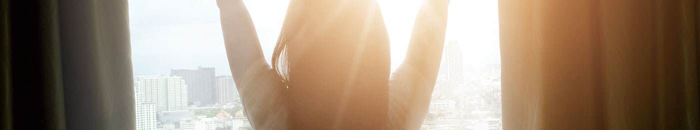 spf boosters -  photo of a white woman opening curtains shown from behind with the light spilling in.
